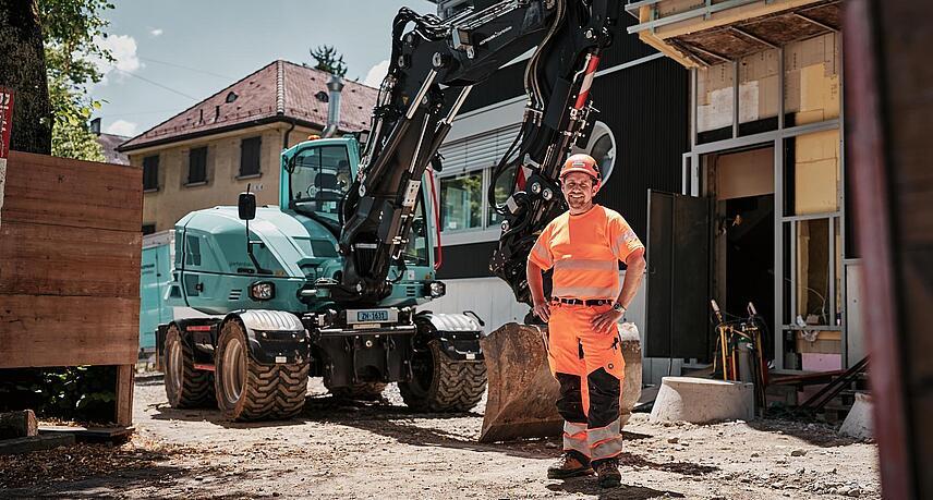 Egy jól látható munkaruhát viselő kezelő egy Wacker Neuson EW100 kerekes kotrógép mellett áll egy városi építkezésen.
