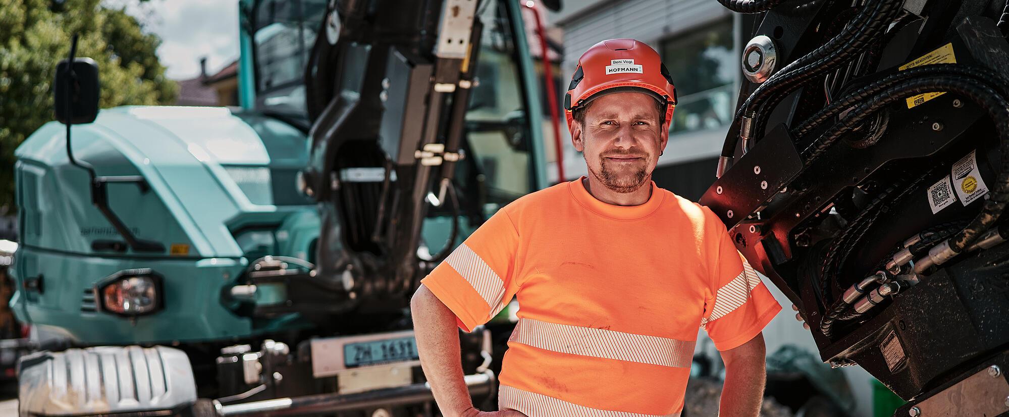 Egy építőipari munkás áll egy hidraulikus mellékberendezéssel felszerelt Wacker Neuson EW100 kerekes kotrógép mellett.
