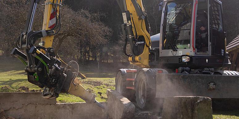 Egy Wacker Neuson EW100 kerekes kotrógép egy erdőtisztáson egy tuskóaprítót működtet, amely egy fatuskót aprít, és faforgácsot szór a levegőbe, miközben a háttérben rönkhalmok és erdő látható.