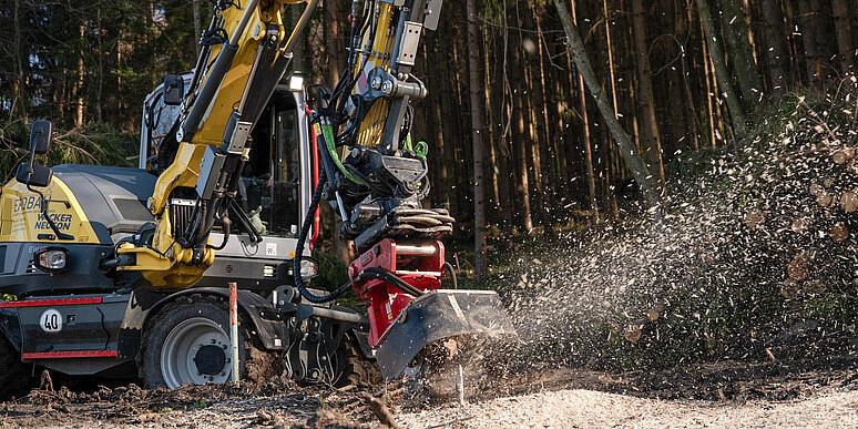 Egy Wacker Neuson EW100 kerekes kotrógép egy erdőtisztáson egy tuskóaprítót működtet, amely egy fatuskót aprít, és faforgácsot szór a levegőbe, miközben a háttérben rönkhalmok és erdő látható.