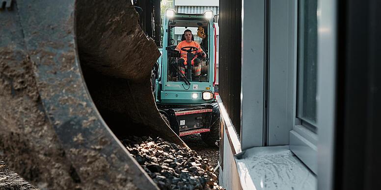 A Wacker Neuson EW100 kotrógép kanállal szállít kavicsot egy épület melletti szűk építési területen.