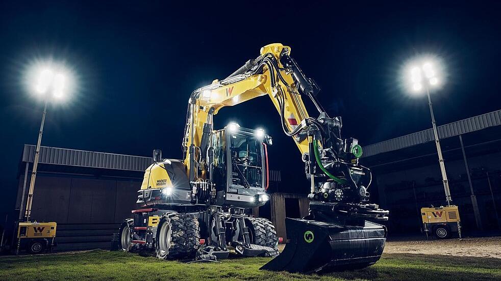 Wacker Neuson EW135 wheeled excavator equipped with a tiltrotator and grading bucket, illuminated under floodlights in a nighttime work environment.