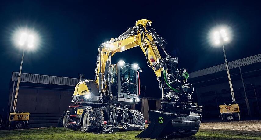 Wacker Neuson EW135 wheeled excavator equipped with a tiltrotator and grading bucket, illuminated under floodlights in a nighttime work environment.