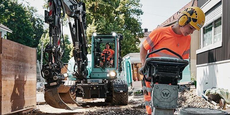 Egy építőmunkás egy Wacker Neuson döngölőt kezel, miközben a háttérben az EW100 kotrógép dolgozik.